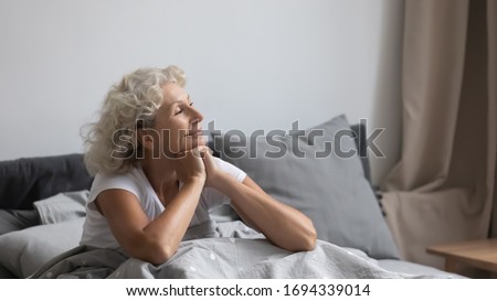Peaceful calm positive middle aged senior retired woman sitting in bed after wakeup in weekend morning, enjoying good mood after good night rest relaxation, welcoming new day alone in bedroom. Royalty-Free Stock Photo #1694339014