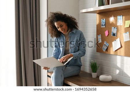 Young happy curly african girl woman lady student blogger freelancer sit table look at modern laptop screen, smiling teen girl type read funny text message email search use application chat online.