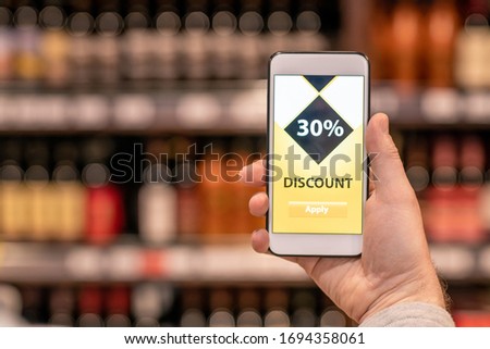 Hand of mobile mature customer holding smartphone with discount application on screen while buying products in supermarket