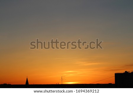 Sun below the horizon and the roof of the church with a cross, television antenna, roof of a multi-storey urban building in the background fiery dramatic orange sky at sunset or dawn backlit by the