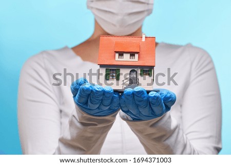 Woman in medical gloves and mask holds house in hands. Concept of home stay, quarantine, security inside the house. Covid-19 Corona Prevention Measures