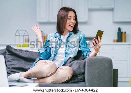 Young attractive woman sitting at home on sofa and showing sincere emotions of excitement about money win in online lottery. Cheerful girl hit the jackpot.