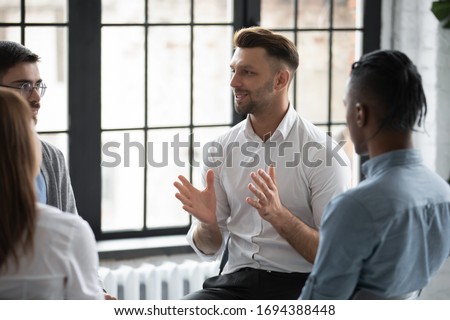 Happy male psychologist counselling speaking with diverse people sitting in circle at group therapy session. Business coach training staff, having fun, team building activity at work. Royalty-Free Stock Photo #1694388448