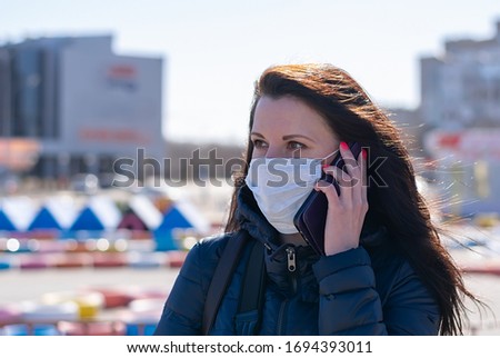 a beautiful girl in a jacket and a medical anti virus mask is talking on the phone in the square against the background of shopping centers and a Playground