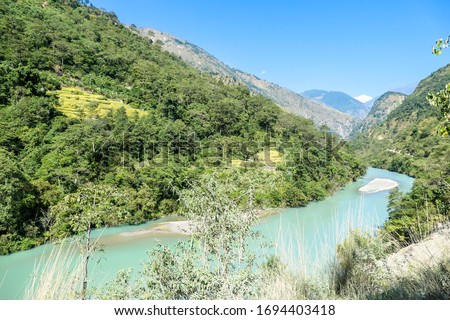 A panoramic view on river and Himalayas from Annapurna Circuit Trek, Nepal. Turquoise color of the river, big stones popping out of the river. Green forest around. Idyllic landscape.