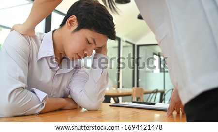 Photo of office worker comforting her colleague. Stressed man in white shirt keeping hand on head while sitting at the wooden working desk over comfortable office as background.