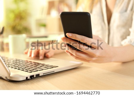 Close up of woman hands checking smart phone using laptop on a desk at home 