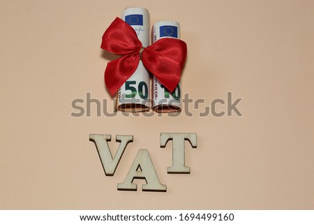 Word VAT made of wooden letters and banknotes of 50 and 10 euro in rolls tied with red ribbon bow top view on pastel color background