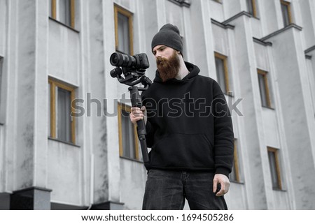 Bearded Professional videographer in black hoodie holding professional camera on 3-axis gimbal stabilizer. Filmmaker making a great video with a professional cinema camera. Cinematographer.