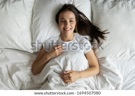 Top view of happy millennial girl lying in white comfortable bed rest on soft fluffy pillow look at camera smiling, overjoyed young woman relax in cozy bedroom wake up in morning feel optimistic