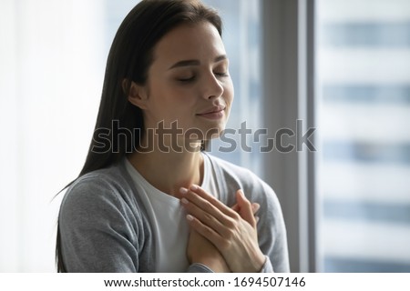 Happy millennial girl hold hands at heart chest feel thankful pray to God at workplace, calm peaceful young woman believer grateful for luck or fate, faith, superstition, gratitude concept Royalty-Free Stock Photo #1694507146
