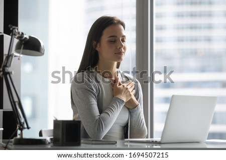 Peaceful young woman worker sit at workplace hold hands at heart chest feel grateful thankful for good luck or fate, millennial female employee believer meditate pray at desk in office, faith concept Royalty-Free Stock Photo #1694507215