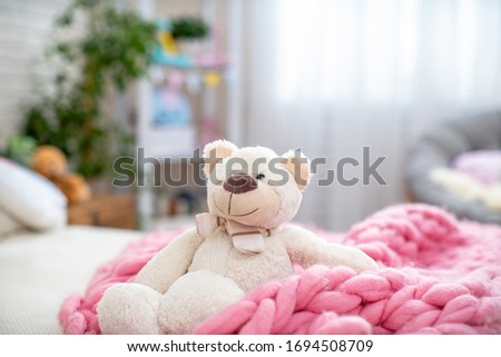 Cute white soft Teddy bear, sitting in a soft Merino blanket, large knitting, on the bed in the children's room. Childhood.