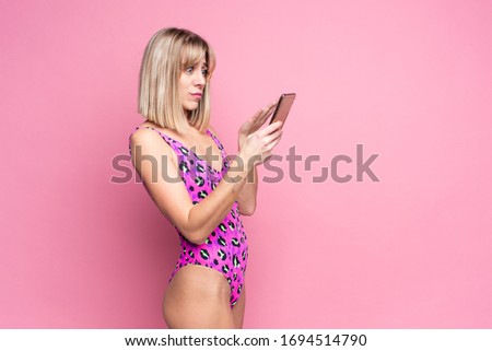 Smiling slim blonde woman in pink leopard swimsuit using smartphone. Standing over pink background, space for text