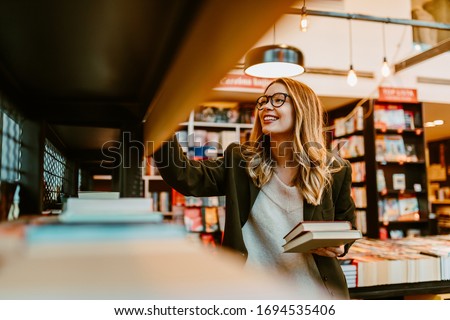 Photo of young blonde woman with glasses looking at book shelves at bookstore. Young woman is looking for new books to buy. Royalty-Free Stock Photo #1694535406