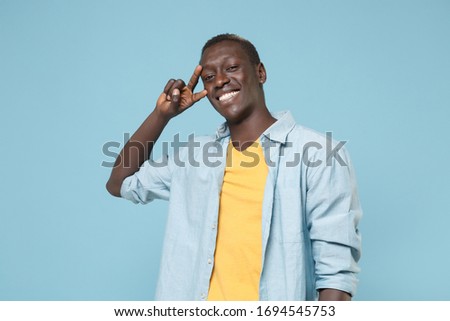 Smiling young african american man guy in casual shirt, yellow t-shirt posing isolated on blue background studio portrait. People emotions lifestyle concept. Mock up copy space. Showing victory sign