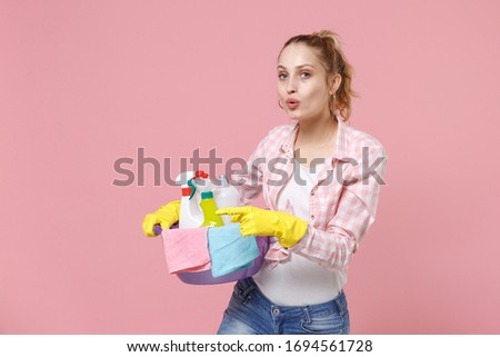 Amazed young woman housewife in rubber gloves hold basin with detergent bottles washing cleansers doing housework isolated on pink background studio. Housekeeping concept. Pointing index finger aside