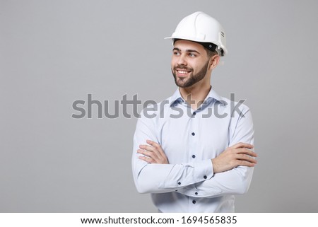 Smiling young unshaven business man in light shirt protective construction helmet isolated on grey wall background. Achievement career wealth business concept. Holding hands crossed, looking aside Royalty-Free Stock Photo #1694565835