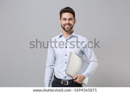 Smiling young unshaven business man in light shirt posing isolated on grey wall background studio portrait. Achievement career wealth business concept. Mock up copy space. Hold laptop pc computer Royalty-Free Stock Photo #1694565871