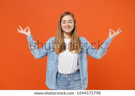 Smiling young woman girl in casual denim clothes posing isolated on orange background. People lifestyle concept. Mock up copy space. Hold hands in yoga gesture relaxing meditating keeping eyes closed