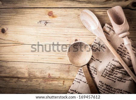 Rustic cooking utensils with a wooden spoon, ladle and scoop lying on a folded cloth with a recipe on it on an old cracked wooden table with vignetting and copyspace