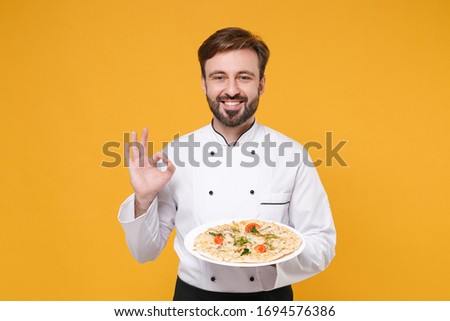 Smiling young bearded male chef cook or baker man in white uniform shirt posing isolated on yellow background. Cooking food concept. Mock up copy space. Hold italian pizza on plate showing OK gesture