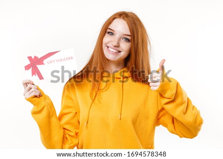 Smiling young redhead woman girl in casual yellow hoodie posing isolated on white wall background studio portrait. People lifestyle concept. Mock up copy space. Hold gift certificate showing thumb up