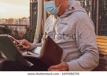 Man with a mask working from home sitting on a wooden bench outdoors with a laptop a telephone and an agenda in a time of confinement due to the covid 19 pandemic