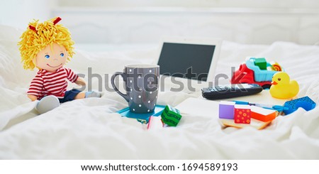 Laptop, cup of coffee, notebook, phone and different toys in bed on clean white linens. Freelance, distance learning or work from home with kids concept