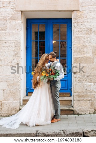 Gorgeous newlyweds with a bouquet in their hands looking at each other standing against the backdrop of a restaurant with blue doors
