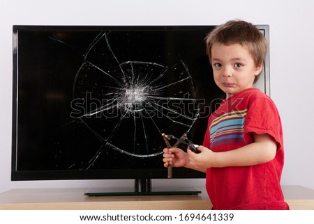 Shocked little boy standing in front of a TV with broken screen holding a slingshot. Home insurance or qarantine problems concept.