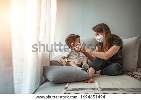 Mother with protective mask holding his son at home do to pandemic outbreak quarantine. Mother putting protective face mask on her child. Side view of adult woman putting on medical mask on little boy