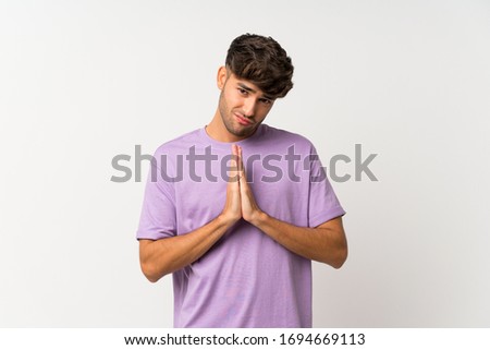 Young handsome man over isolated white background keeps palm together. Person asks for something