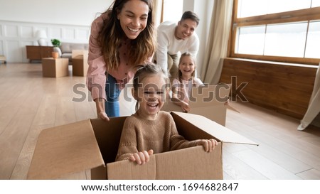 Happy little girls sisters sitting in big carton boxes while joyful parents pushing it on floor. Laughing family couple having fun with small children in new house, relocation moving day concept. Royalty-Free Stock Photo #1694682847