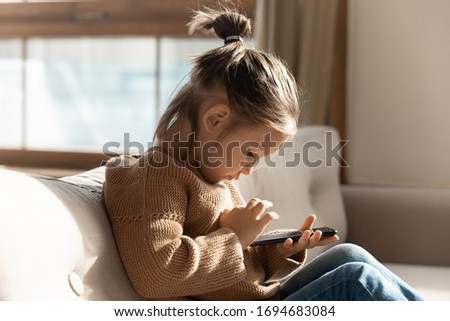 Side view little preschool girl sitting on sofa, using smartphone applications alone in living room. Addicted to technology small child playing online mobile games, web surfing internet at home.