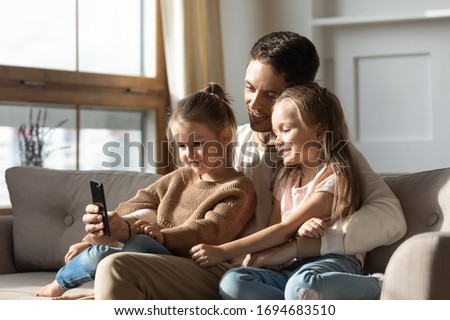 Happy young father cuddling two little daughters, showing funny cartoons video on smartphone. Smiling single dad embracing small children, taking selfie, relaxing together on comfortable sofa.