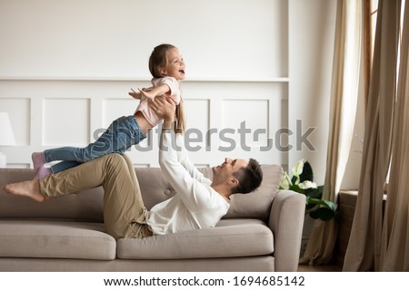 Side view overjoyed little girl playing plane with happy young daddy on couch. Cheerful dad lying on sofa, holding lifting on straight hands small playful daughter, having fun together in living room. Royalty-Free Stock Photo #1694685142
