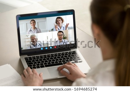 View over client shoulder sit at desk receive medical consultation on-line from diverse specialists. Woman listen doctors about corona virus precautionary measures, videoconference laptop webcam view