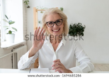 Close up headshot portrait picture of happy 60 years old businesswoman sitting by table. Smiling attractive young mature woman mentor greeting looking at camera in office.