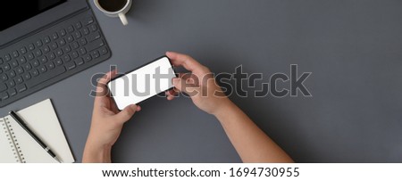 Overhead shot of businessman using horizontal mock-up smartphone while sitting at workspace with notebook and digital devices on grey table