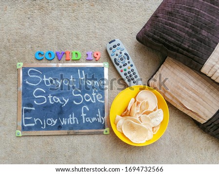 Flat lay, a top view of a massage board with text stay at home stay safe with a remote control, some snack and cushions. Pandemic Covid-19 Corona virus quarantine concept. 