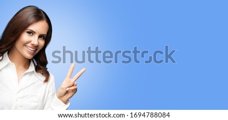 Portrait of happy smiling young cheerful confident businesswoman, showing two fingers or victory hand gesture, over light blue background. Success in business concept picture. 