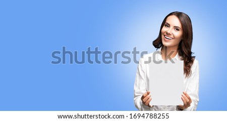 Portrait of happy smiling brunette businesswoman showing blank signboard with copy space area for some slogan or text, over light blue color background. Success in business concept. 