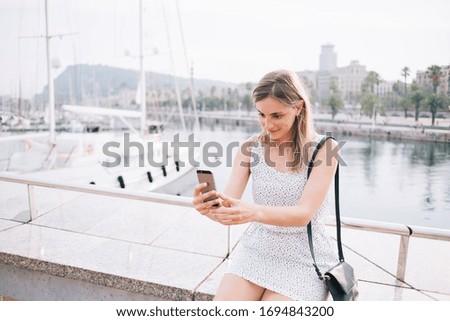 Satisfied female tourist taking selfie for travel blog on social media while sitting on stone parapet in port on background of waterfront