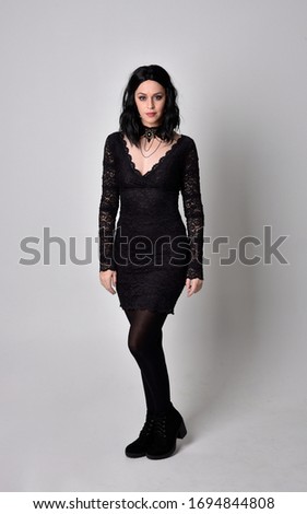 Portrait of a goth girl with dark hair wearing black dress and boots. Full length standing pose on a studio background.