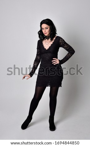 Portrait of a goth girl with dark hair wearing black dress and boots. Full length standing pose on a studio background.