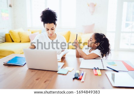 African American mother checking notes in school notepad with playful little girl using phone and picturing mother distracting from homework 