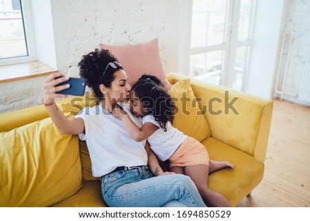 From above black young mother with closed eyes kissing cute little girl in forehead and taking selfie on couch at home