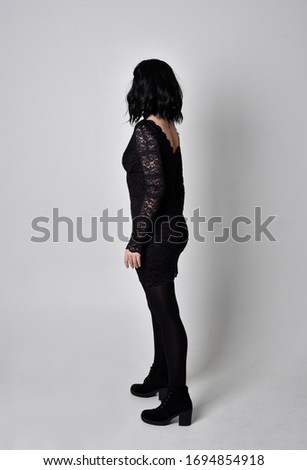 Portrait of a goth girl with dark hair wearing black and plaid skirt with boots. Full length standing pose with back to the camera on a studio background.