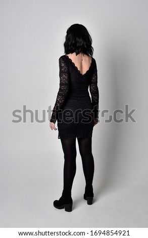 Portrait of a goth girl with dark hair wearing black and plaid skirt with boots. Full length standing pose with back to the camera on a studio background.